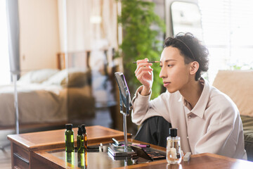自宅リビングでメイクアップをしている若い日本人男性/ペンシルで眉を描いている/メンズビューティー