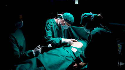 Doctor takes surgical knife from an assistant's hand operate on patient lying on bed, bent down skillfully cut patient's skin with his knife, assistant prepares a quick surgical swab for doctor.