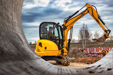 Mini excavator at the construction site on the edge of a pit against a cloudy blue sky. Compact...