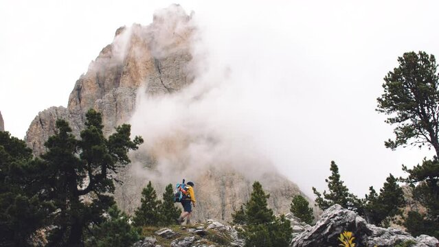 Young man hiker hiking on stone mountain with cloudy sky and fog. Yellow jacket, backpack, black beard and beanie. Traveling Dolomites, Italy.