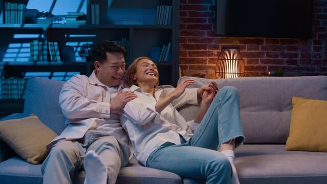Happy multiracial diverse couple Asian man and Caucasian woman homeowners renters jumping on sofa in new apartment relax on couch at night evening laughing hugging real estate mortgage loan tenancy