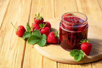Strawberry jam in glass jar on wooden board with fresh strawberry fruit and green leaves on wooden background. Recipe of delicious homemade berry jam of strawberry full of vitamins and antioxidants.