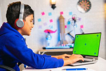 Young indian teenager kid with headphones working on green screen laptop at home - concept of technology, brilliant and e-learning