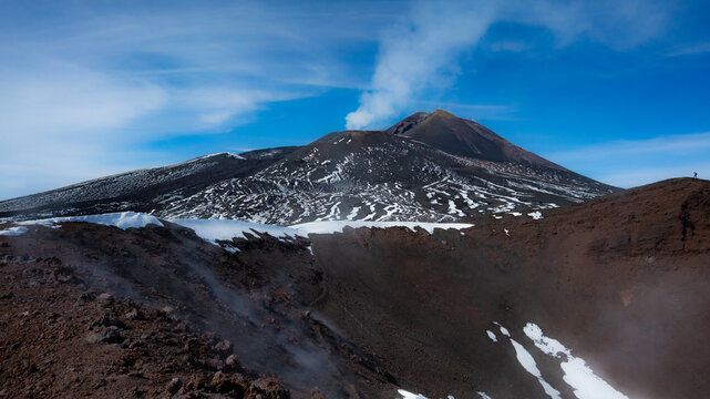 Majestic scenery of Enta (Sicily, Italy) main craters with smoke and gases coming out, a hiking adventure on volcano above the clouds in early spring with patches of snow, blue sky