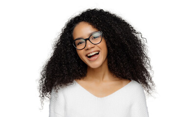 Carefree overjoyed dark skinned woman laughs happily, being in high spirit, tilts head, has curly dark hair, wears white jumper, hears something funny, models against grey studio background.