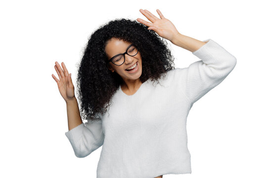 People and joy concept. Optimistic delighted Afro American woman raises hands, dances to loud music, being overjoyed, wears casual white jumper, isolated against blue background, has good mood