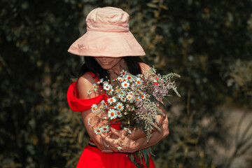 A girl in a pink hat, glasses in a red dress holds a bouquet of wild flowers in her hands on a blurred background. Summer outdoor recreation. Close-up.