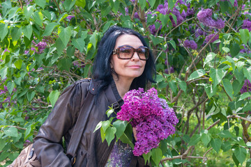 A happy girl with long black hair in glasses smells the scent of lilac flowers in the garden. Fragrant lilac flowers. Blooming lilac bushes in the garden. Rest in the botanical garden.