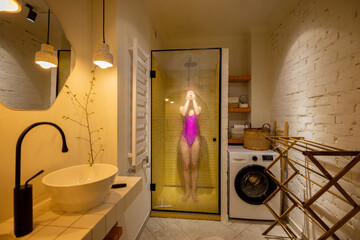 Woman in pink swimsuit takes a shower in the shower cabin at home. Wide interior view of stylish white bathroom. Concept of everyday routine, hygien and domestic lifestyle