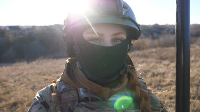 Close up blue eyes of female ukrainian army soldier looking into camera. Sight of young military woman in helmet and balaclava at countryside. Invasion of territory Ukraine. End of war in Europe