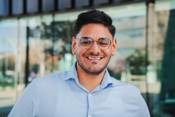 close up portrait of hispanic young entrepreneur smiling and looking at camera at workspace. Front view of one latin happy employee office worker with glasses and successful attitude standing outdoors