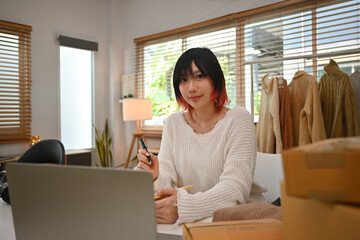 Stylish young female small business owner sitting at working desk with cardboard boxes and laptop