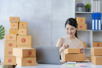 Charming asian woman starting a small business, SME entrepreneur, distribution warehouse with post box. owner's small home office Online marketing and product packaging and home delivery services.
