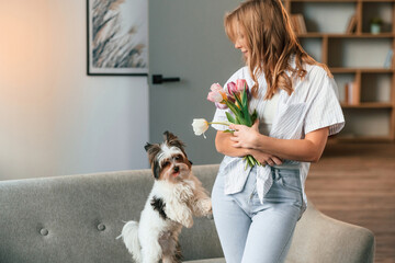 With flowers in hands. Beautiful young woman is with little dog indoors