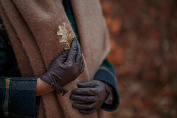 Female hands in brown leather gloves with a dried oak leaf against the background of a terracotta woolen scarf and a plaid coat