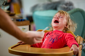 crying and angry little girl sitting in a high chair and refusing a bowl of food, period of...