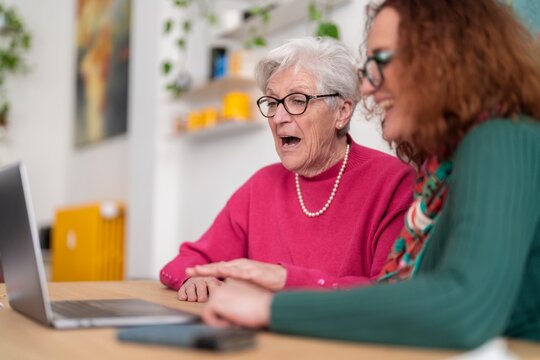 Soft focus of cheerful elderly gray haired female looking at screen while using computer with redhead daughter sitting at table