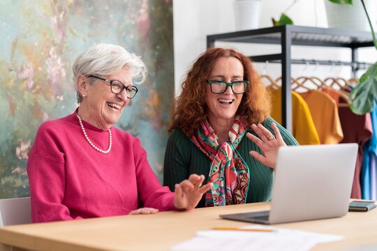 Cheerful elderly gray haired female in glasses clapping hands while sitting with mature daughter waving hand during video call on computer