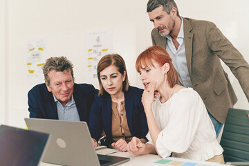 Team Project Review - A diverse group of four junior and senior colleagues gather around a laptop, studying a project together with focused expressions, dressed in casual attire.