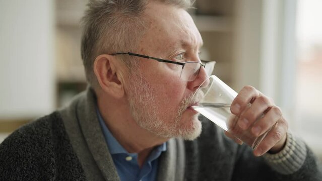 Portrait of thirsty gray haired senior man drinking clear water from glass. Elderly male in sunglasses taking care health. Maintaining water balance in human body and healthy good life habits concept.