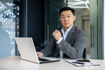 Serious thinking asian businessman working inside office sitting at table with laptop, boss making important financial decision, asian mature man in business suit at workplace.