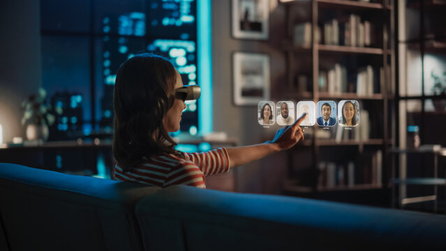 Young Woman Using Virtual Reality Headset At Home, Sitting On Couch In Living Room, Making Gestures To Answer AR Video Call Chat With Family Doctor. Medical Health Online Examination And Doctor Visit.