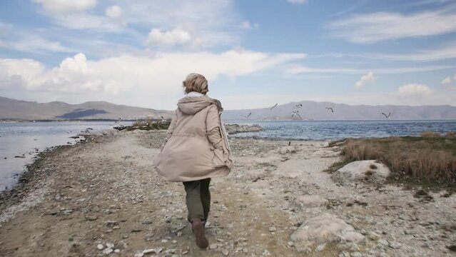 A cheerful young adult woman in a jacket runs along the shore of a mountain lake and frightening sea gulls the birds take off in slow motion. Feeling of freedom and joy in life
