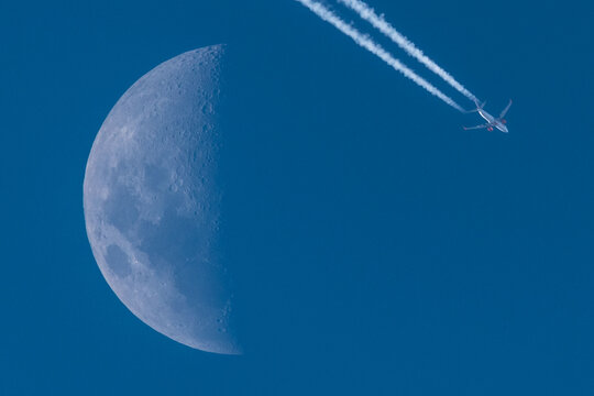 avion de aerolinea dejando una estela blanca pasando a la derecha de la luna en fase cuarto creciente durante el dia