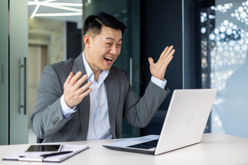 Successful asian investor financier celebrating victory and triumph, mature boss in business suit working inside office with laptop, man received online win news.