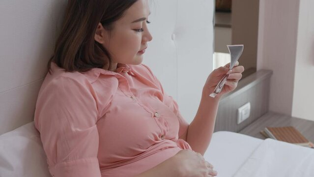 Happy young pregnant woman looking at her baby ultrasound picture while laying in her bed in her bedroom and showing it to the camera