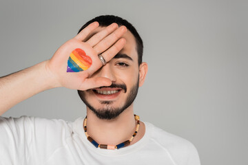Smiling gay man covering eye with lgbt flag on hand isolated on grey, International day against...