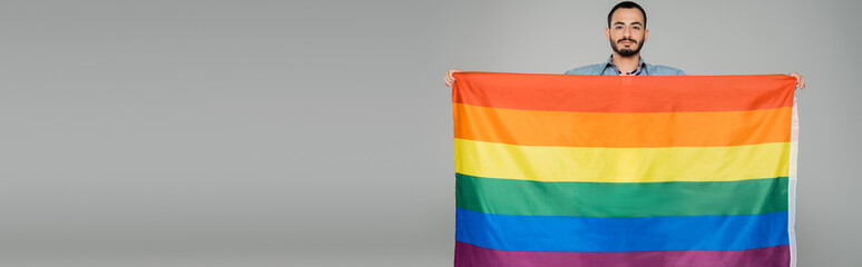 Brunette gay man holding lgbt flag and looking at camera isolated on grey, banner.
