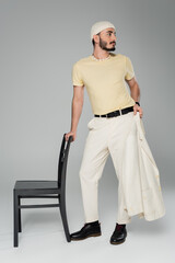 Full length of well dressed homosexual man holding jacket near chair on grey background.