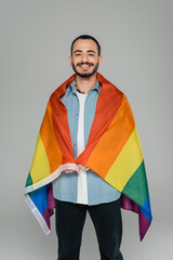 Cheerful gay man looking at camera and holding lgbt flag isolated on grey, International Day...