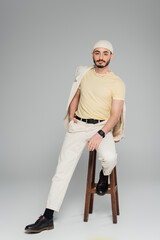 Well dressed gay man in suit and hat posing on chair on grey background.