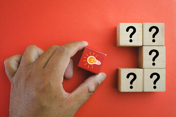 Hand Choosing Red wooden cube with glowing light bulb icon among question mark icon on wooden cubes over red background use for Creativity and inspiration ideas,Business idea,Thinking concept.