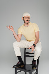 Confused gay man in hat sitting on chair isolated on grey.