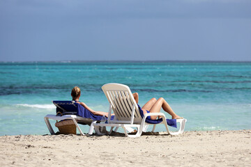Two girls in bikini tanning on white deck chairs on sea waves background. Vacation on sandy beach