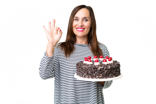 Middle age caucasian woman holding birthday cake over isolated chroma key background showing ok sign with fingers
