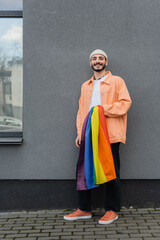 Smiling gay man holding lgbt flag near building outdoors.