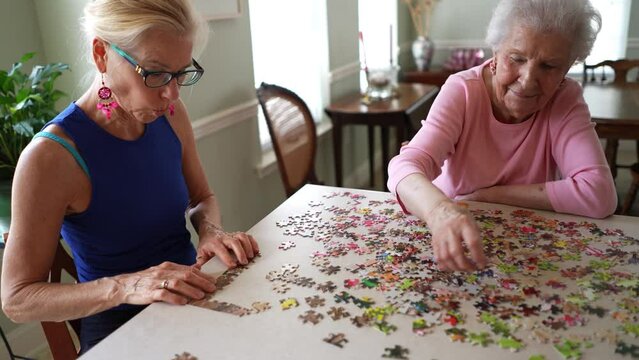 Camera turns around two happy elderly mature women talking and playing with puzzle pieces.
