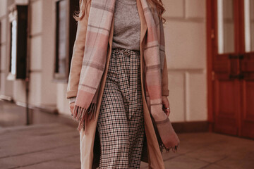 Female figure wearing beige long autumn warm coat, checkered pants and scarf. Outdoor street style fashion portrait