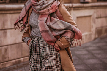 Woman in autumn stylish fashion brown long coat, scarf and plaid pants walking in the city. Female casual street style outfit - obrazy, fototapety, plakaty