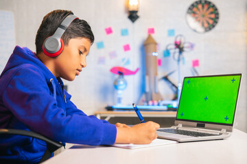 Indian kid making notes from online class on green screen laptop at home - concept of e-learning, intelligence and education
