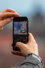 Ein in der Hand gehaltenes Mobiltelefon welches gerade ein Panoramabild von einer Stadt mit wolkigen Himmel in Deutschland aufnimmt. 
