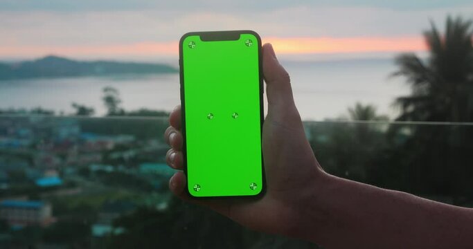 Holding mobile phone with green screen chroma key over sunset at sea at tropical scenery. Male hand with mobile phone with green screen mockup on tropical beach at sunset on summer vacations