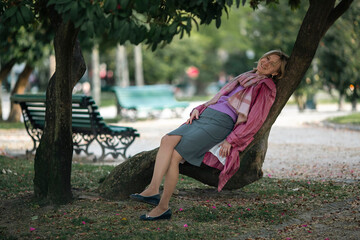 A woman is resting on the roots of a tree in the park.