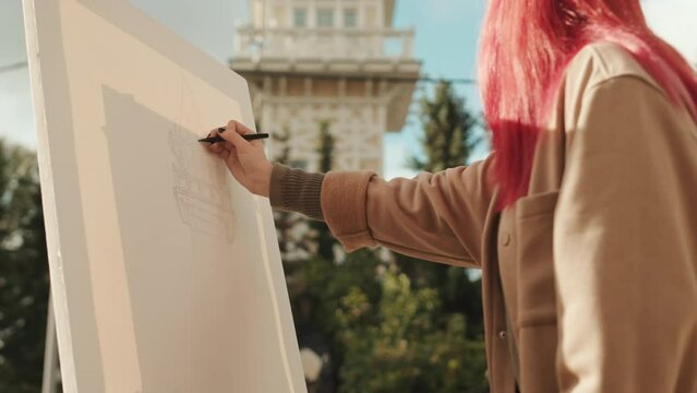 Young female artist with sunlit red hair drawing beautiful castle on canvas outdoors in summer