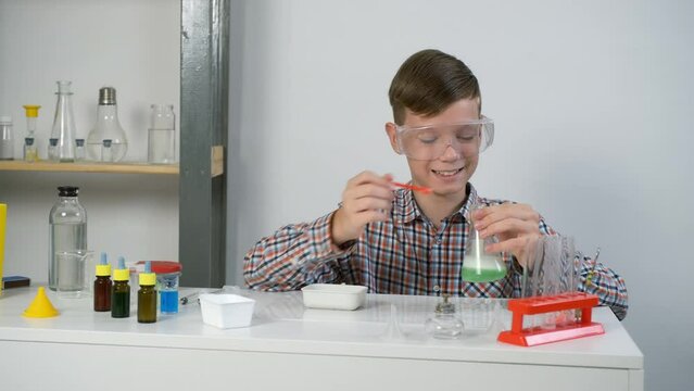 Teen boy is wearing protective glasses and doing chemistry experiments with gas reaction. He is mixing soda, citric acid and green liquid in flask. Online education, homeschooling and science concept.