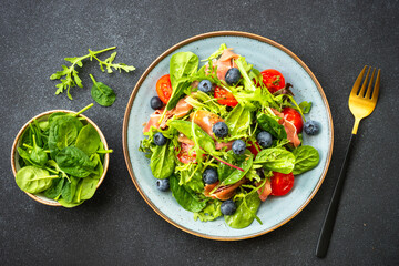 Green salad with fresh prosciutto, green leaves mix and tomatoes at dark table. Top view with copy space.